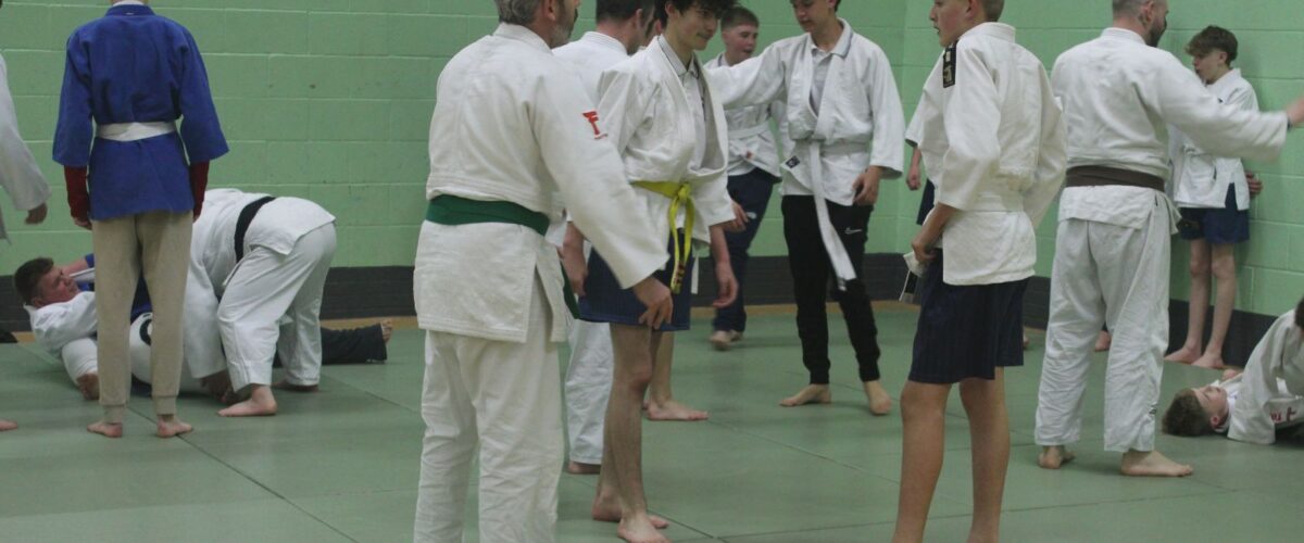 Secondary school pupils taking part in judo. Two boys listening to teacher explain with other pupils stood around them in sports hall.